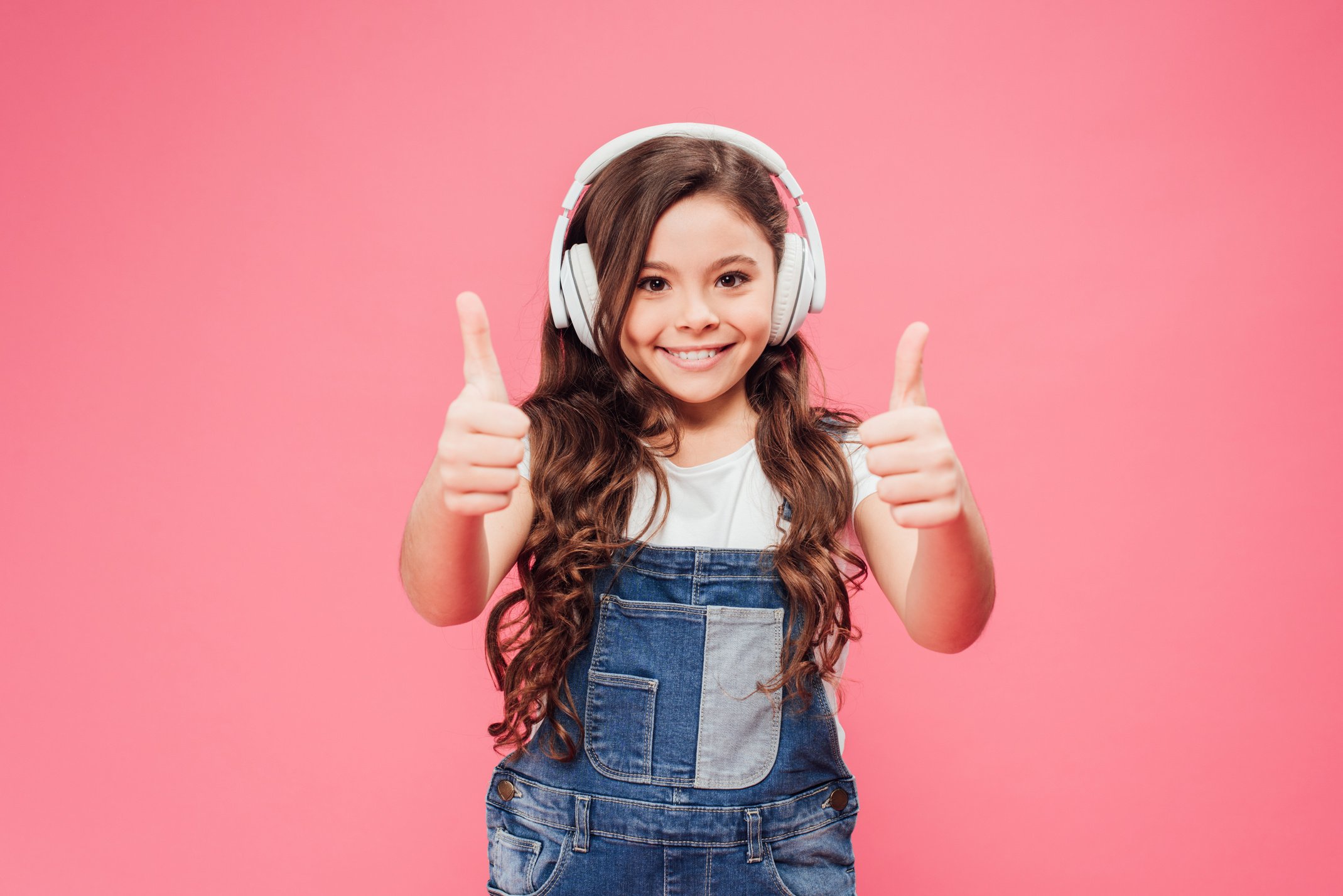 smiling kid in headphones showing thumbs up isolated on pink